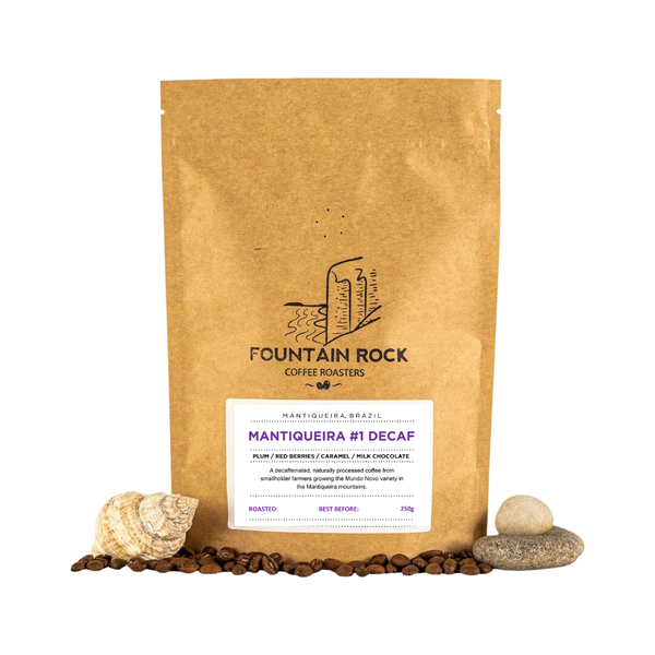 A brown paper coffee bag labeled Fountain Rock Coffee Roasters Mantiqueira #1 Decaf stands upright on a white background, with coffee beans and small stones arranged around the base.